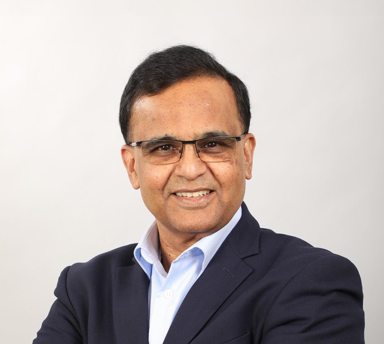 A man in a dark blazer and rectangular glasses smiles against a gray background, his short, dark hair brushed to the side.